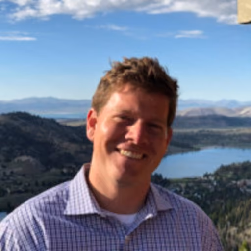 Man smiling in a checkered shirt with mountains and lake in the background on a sunny day.