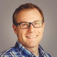 Smiling man with short light brown hair wearing glasses and a blue plaid shirt against gray background.