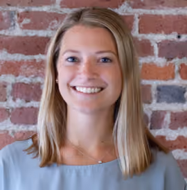 Smiling woman with blonde hair wearing a light blue top standing against a brick wall.