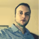Man with short hair and beard wearing a checkered shirt against a blurry indoor background.
