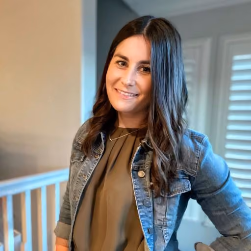 Smiling woman with long dark hair wearing a denim jacket and olive green top indoors near window blinds.