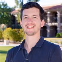 Smiling man with short dark hair wearing a dark polo shirt outdoors near greenery and a building.