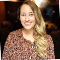 Smiling woman with long blonde hair wearing a brown patterned top against a dark blurred background.