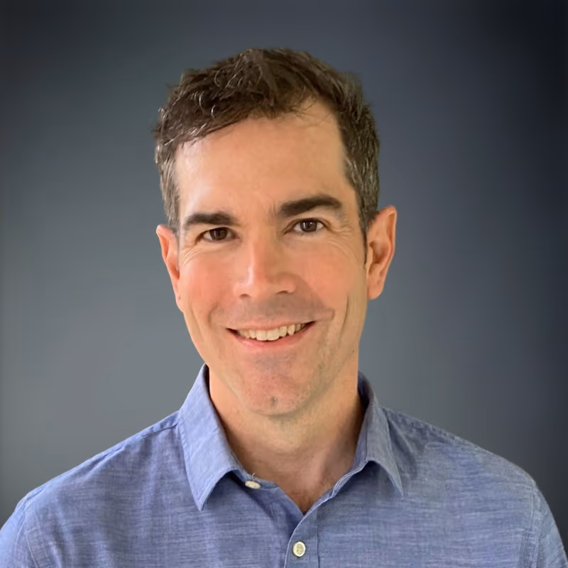 Smiling man with short dark hair wearing a light blue button-up shirt against gray background.