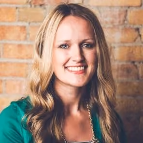 Smiling woman with long wavy blonde hair wearing a green top in front of a brick wall.