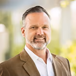 Smiling middle-aged man with beard wearing a brown blazer and white shirt against a blurred background.