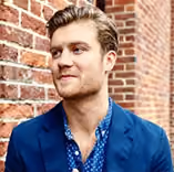 Man with blond hair wearing blue blazer and patterned shirt standing by a brick wall.