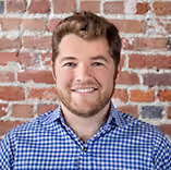 Smiling man with short brown hair in a blue checkered shirt against a brick wall background.
