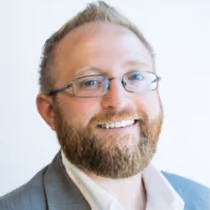 Smiling man with glasses, beard, and suit jacket against a light background.
