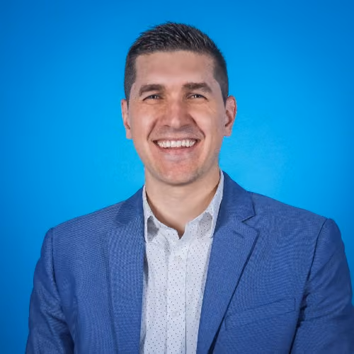Smiling man with short dark hair in a blue suit jacket and white shirt on a blue background.