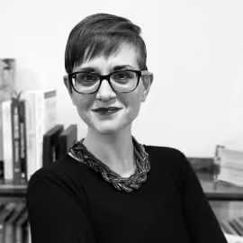 Portrait of a person with short hair, glasses, and a braided necklace smiling in front of bookshelves.