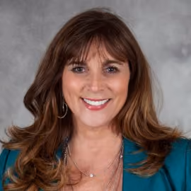 Portrait of a smiling woman with brown hair wearing a teal blazer and earrings against a gray background.