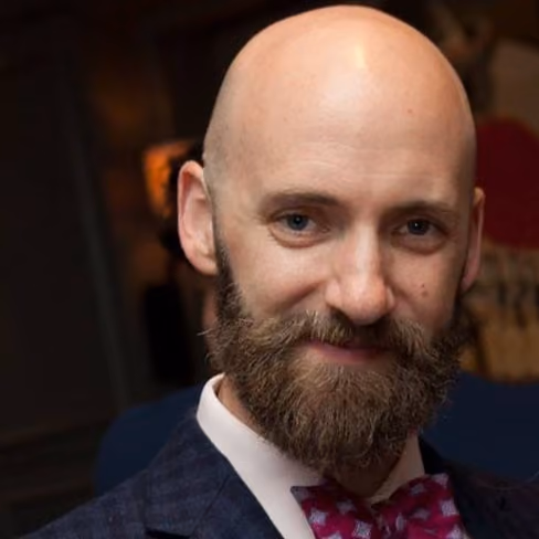 Smiling bald man with a full brown beard wearing a suit and patterned bow tie in a dim room.
