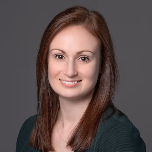 Smiling woman with straight brown hair wearing a dark green top against a gray background.