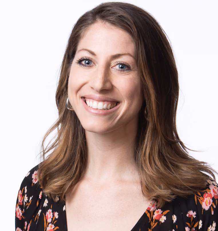 Smiling woman with light brown hair wearing a black floral top against a white background.