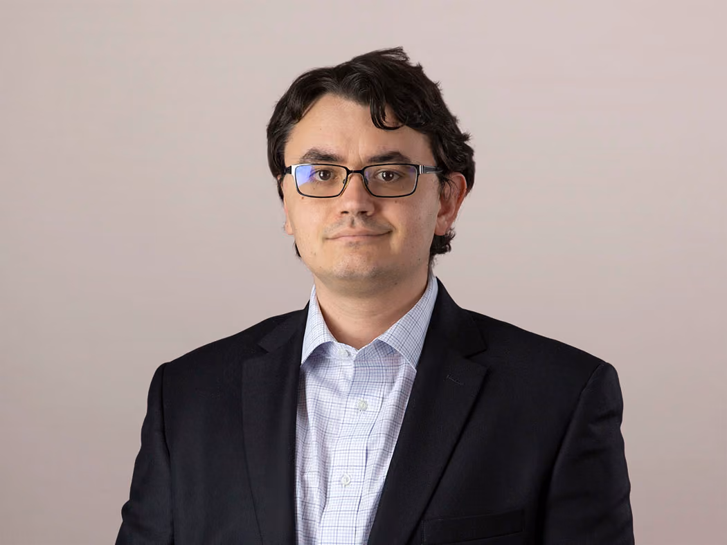Man with dark hair and glasses wearing a black blazer and checked shirt against a plain background.