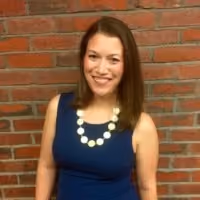 Smiling woman in a blue sleeveless dress wearing a white circular necklace against a brick wall.