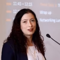 Woman with curly dark hair speaking at a microphone in front of a presentation screen.