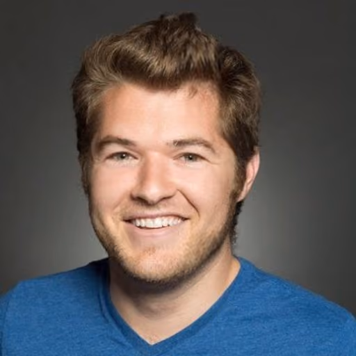 Smiling young man with brown hair wearing a blue shirt against a gray background.