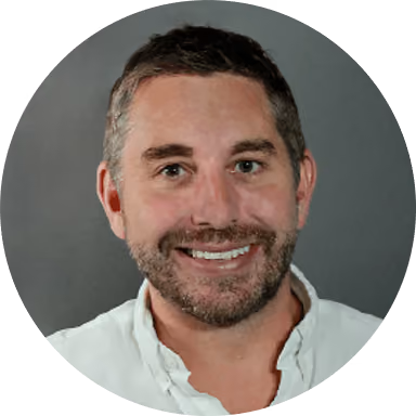 Smiling man with short hair and beard wearing a white shirt against a gray background.
