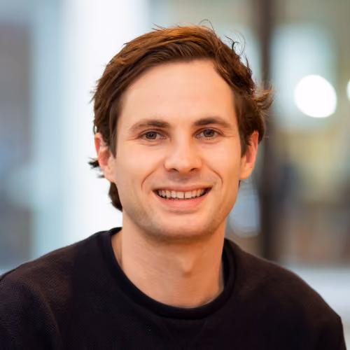 Smiling man with brown hair wearing a dark sweater against a blurred background.