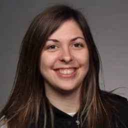 Smiling woman with long brown hair wearing a black top against a gray background.