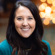 Smiling woman with long dark hair wearing a teal top and black turtleneck in front of warm bokeh lights.