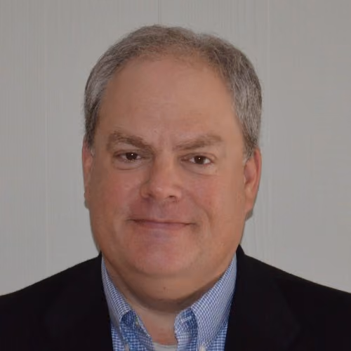 Middle-aged man with short gray hair wearing a blue checkered shirt and dark blazer against a plain background.