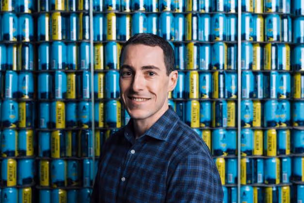 Man in a blue checkered shirt smiling in front of stacked blue and yellow beverage cans.