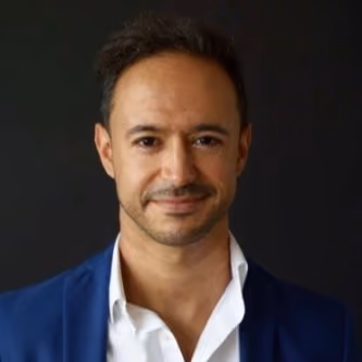 Man with short dark hair and trimmed beard wearing a white shirt and blue blazer against a black background.
