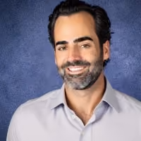 Smiling man with dark hair and beard wearing a light gray collared shirt against a blue textured background.