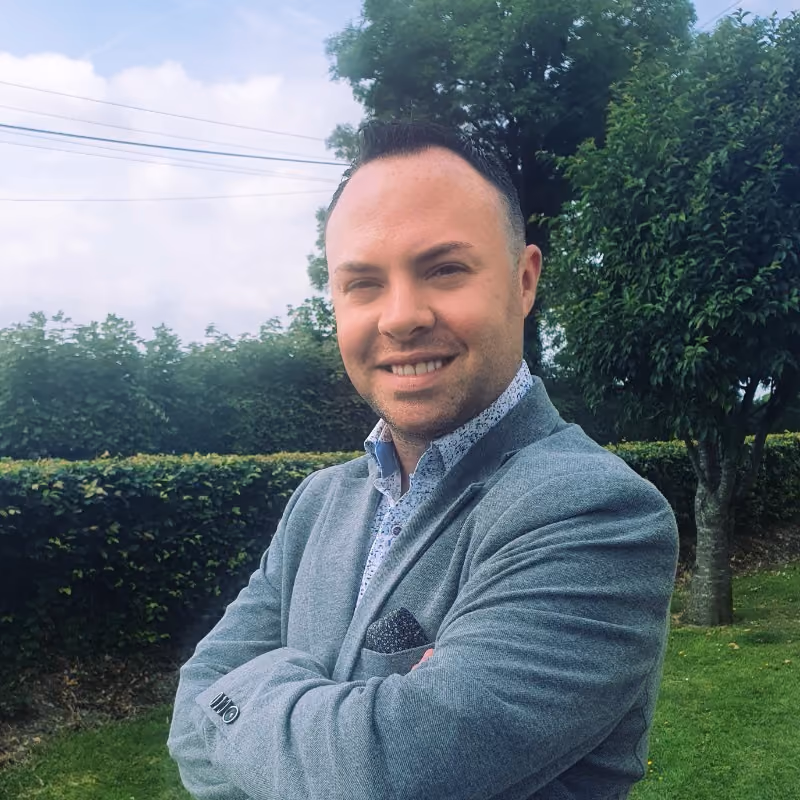 Smiling man in a gray blazer with folded arms standing in a garden with trees and hedges.