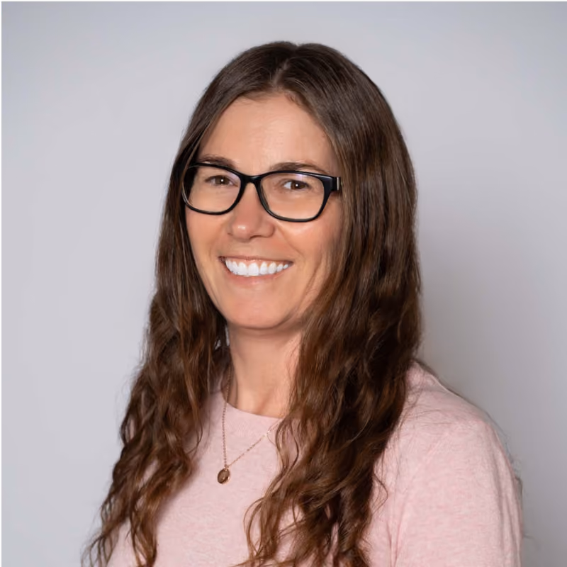 Smiling woman with long wavy brown hair, black glasses, pink top, and gold necklace on a gray background.
