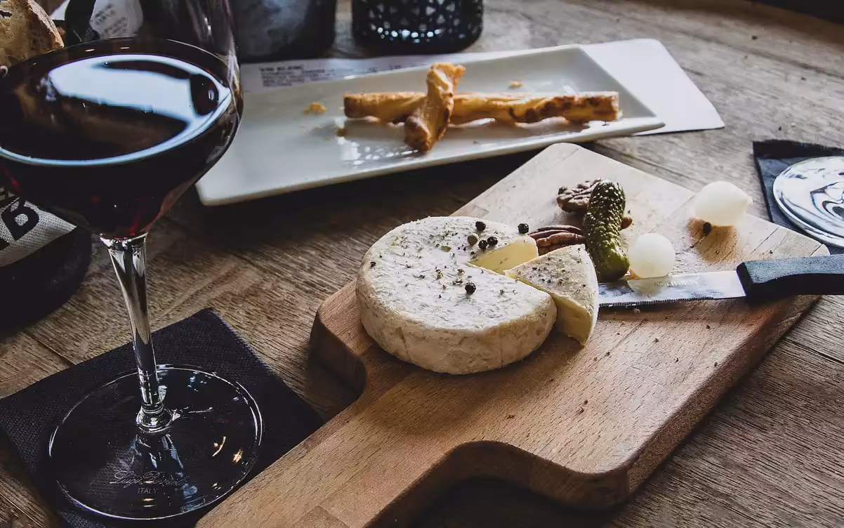 Glass of red wine and a wooden board with a round cheese, pickles, nuts, and a knife on a wooden table.