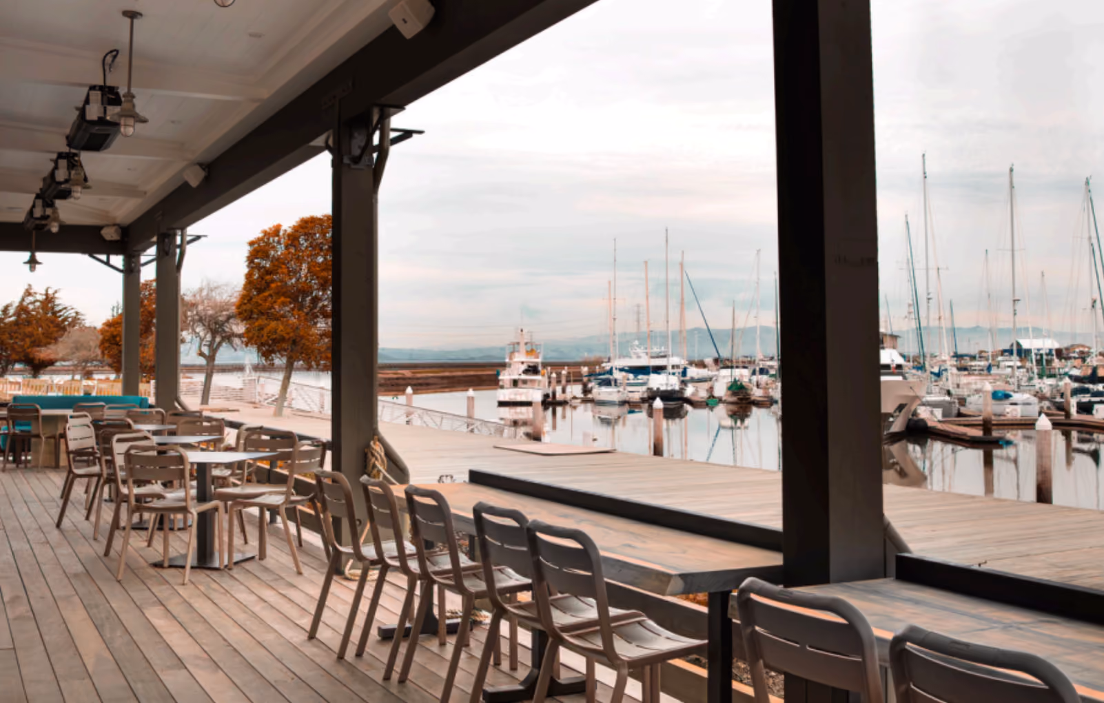 Outdoor seating area with tables and chairs overlooking a marina with sailboats docked.