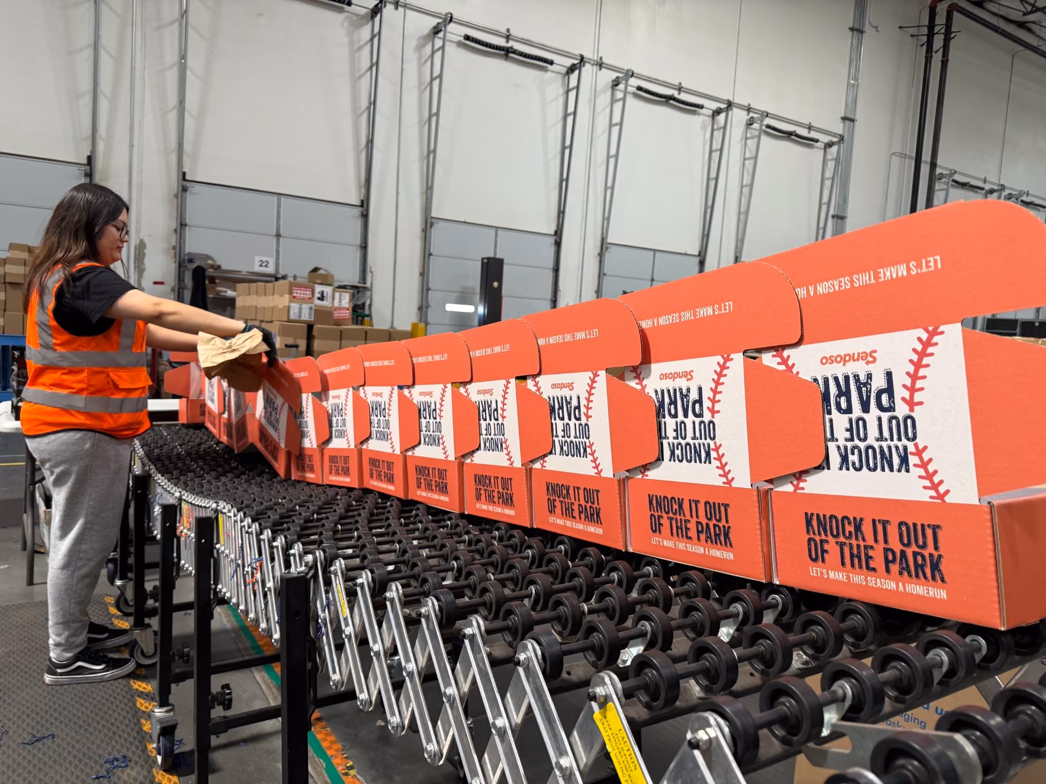 Worker in orange safety vest assembling red 'Knock It Out Of The Park' boxes on conveyor in warehouse.