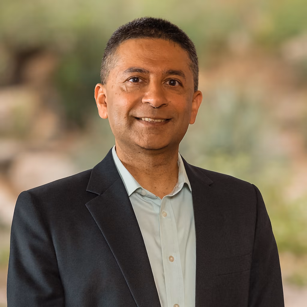 Smiling man in black blazer and light blue shirt with blurred green outdoor background.