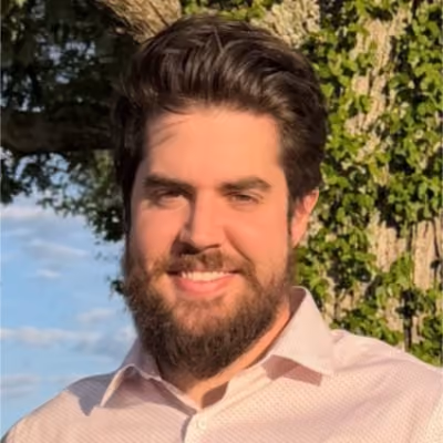 Smiling man with dark hair and beard wearing a light pink button-up shirt outdoors in sunlight.