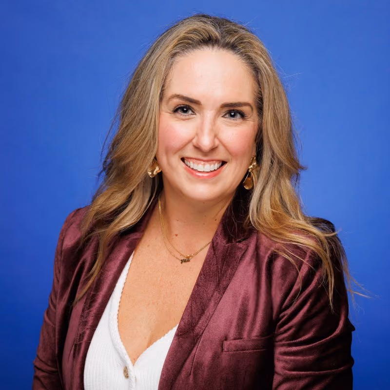Smiling woman with long blonde hair wearing maroon blazer and white top against blue background.