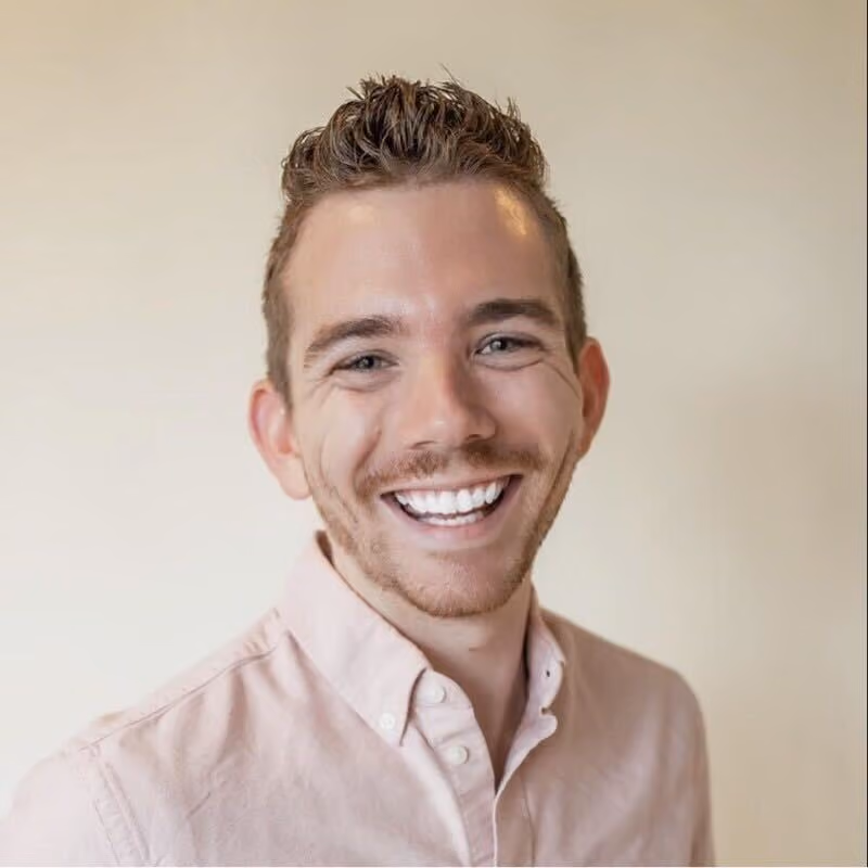 Smiling young man with short curly hair wearing a light pink button-up shirt on a beige background.