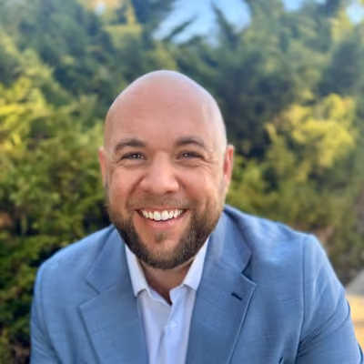 Smiling bald man with beard wearing a light blue suit jacket outdoors with green trees behind him.
