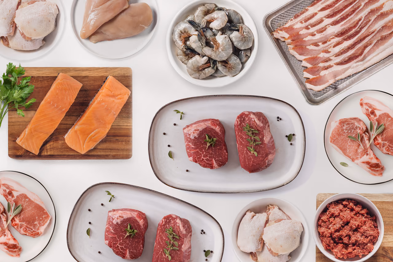Various raw meats including steaks, bacon, salmon, pork chops, shrimp, ground beef, and chicken on plates and cutting boards.