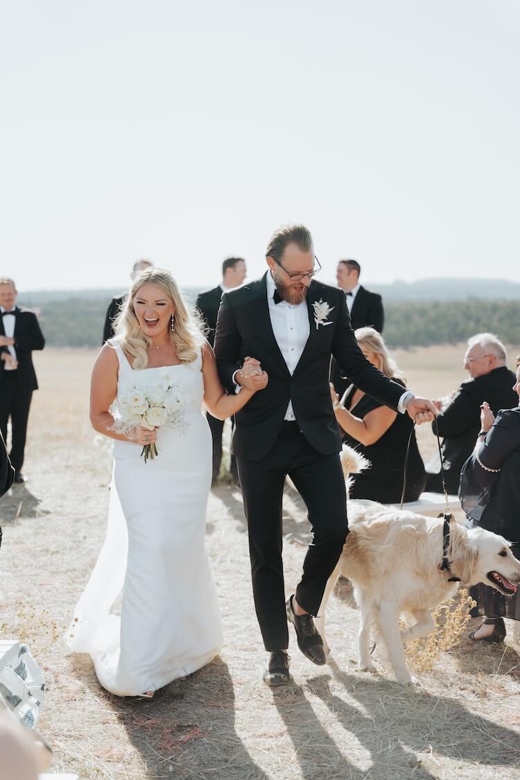 Bride and groom walking down outdoor micro wedding aisle with their dog