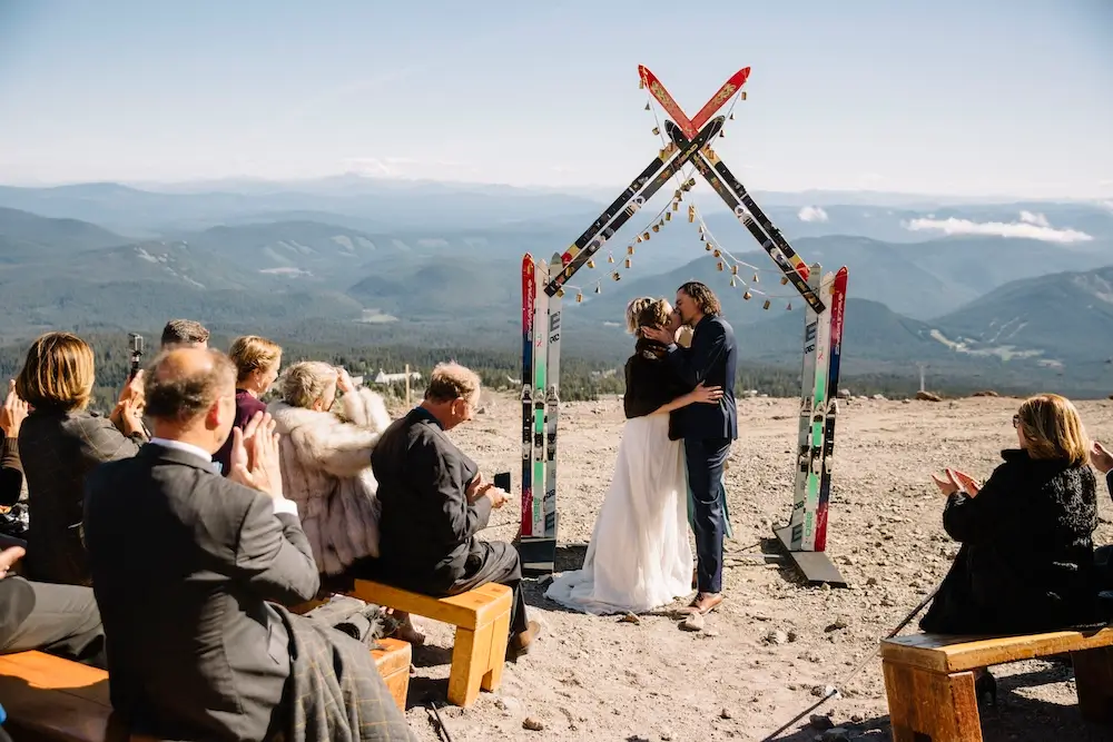 Mountain-top micro wedding ceremony with couple kissing under a unique ski arch, surrounded by family and friends with panoramic alpine views.