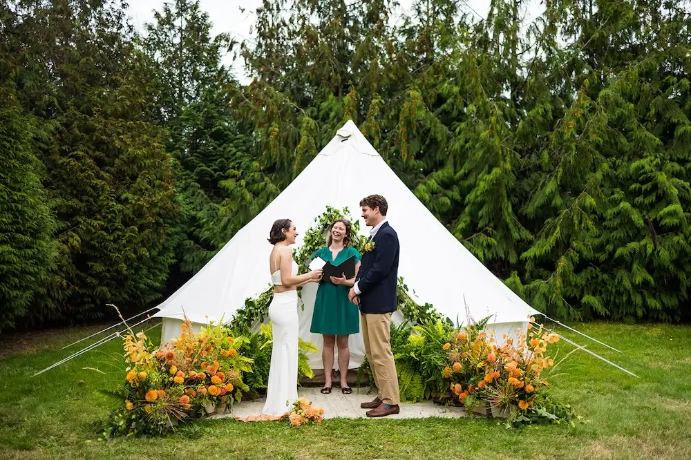 Outdoor micro wedding elopement with couple exchanging vows in front of a white tent, surrounded by lush greenery and vibrant floral arrangements.