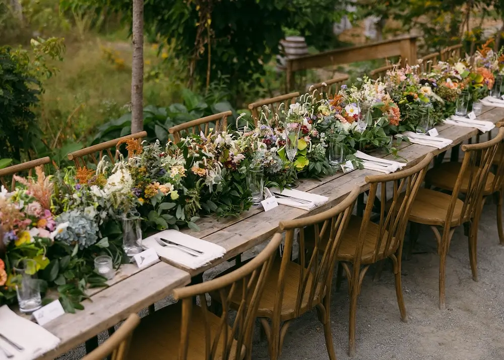 Long wooden table set for an intimate brunch wedding reception with lush garden-style floral arrangements and natural wood chairs in an outdoor setting.