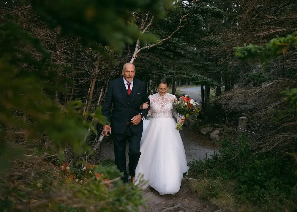 Bride walking with her father along a forest path while holding a bouquet.