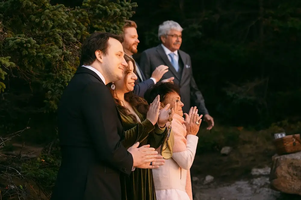 Guests standing outdoors and clapping during a wedding ceremony in warm sunrise light.