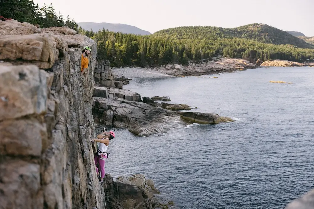 Person rock climbing on a coastal cliff above the ocean with forested shoreline in the background.