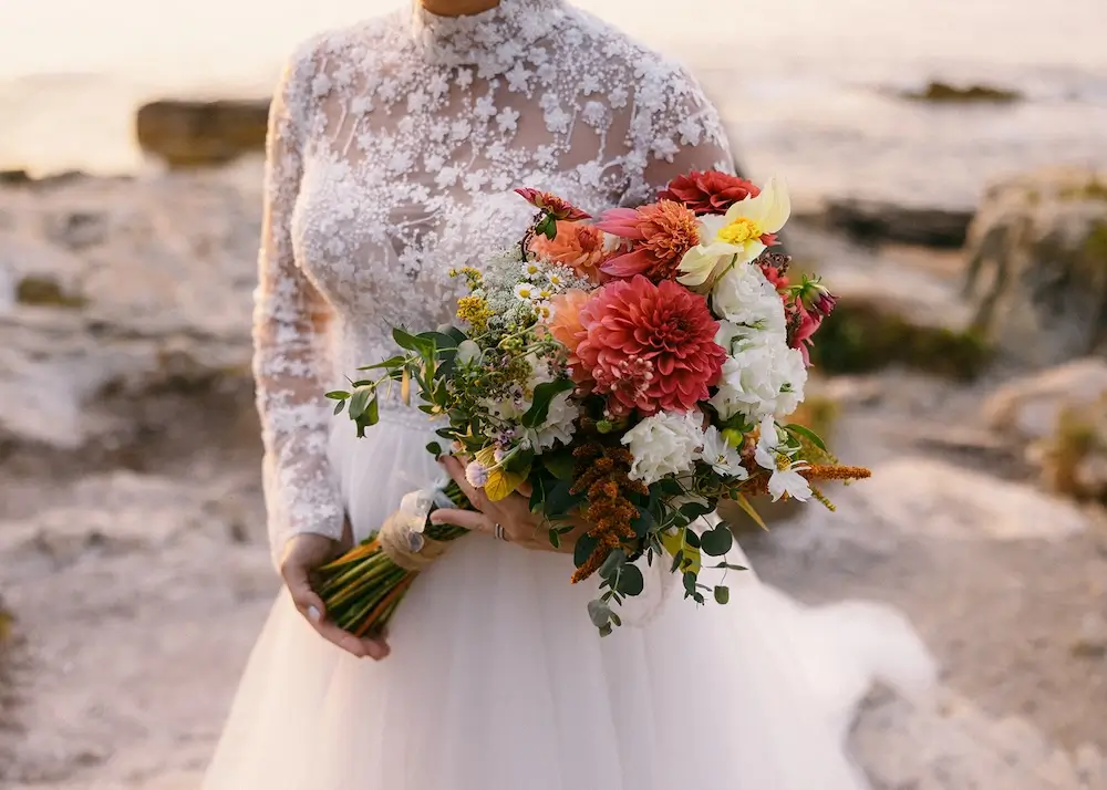 Close-up of a bride holding a colorful bouquet of flowers while wearing a lace wedding dress near a rocky coastline.